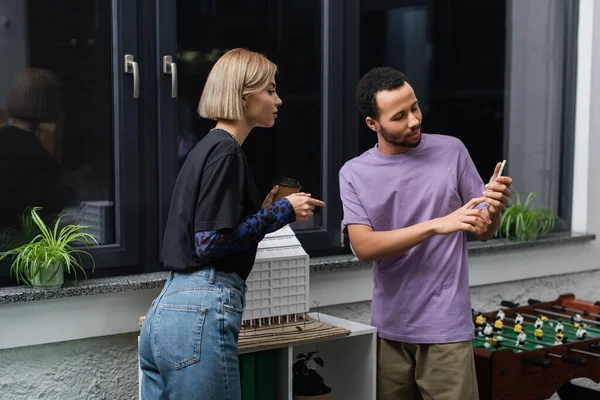 Mujer rubia apuntando a un teléfono inteligente cerca de un colega afroamericano - foto de stock