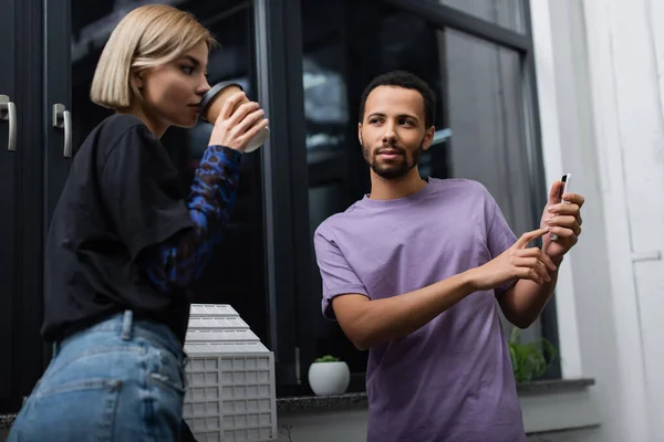 African american man showing smartphone to blonde colleague drinking coffee to go — Stock Photo