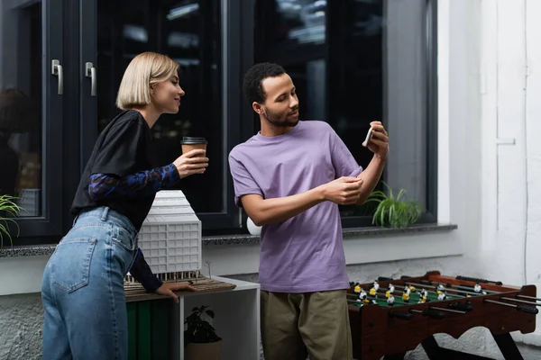 African american man showing smartphone to cheerful colleague holding coffee to go — Stock Photo