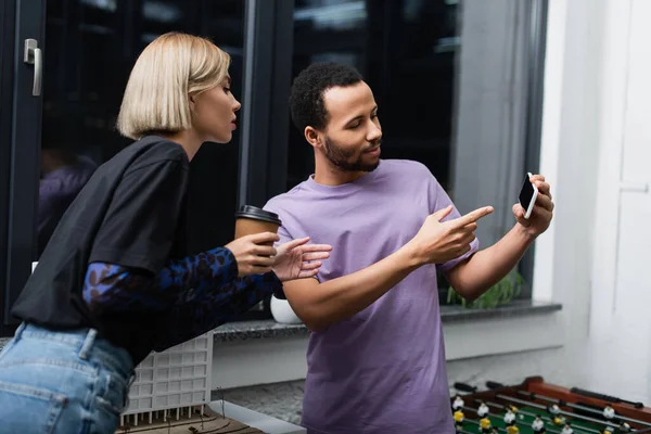 Uomo afroamericano indicando smartphone a collega bionda che tiene il caffè per andare — Foto stock