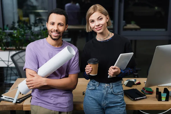 Mujer rubia sosteniendo una tableta digital y una taza de papel cerca de un colega afroamericano con papel enrollado - foto de stock