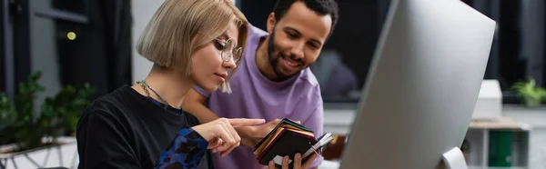Blonde Innenarchitektin in Brille zeigt auf Muster in der Nähe von afrikanisch-amerikanischem Kollegen, Banner — Stockfoto