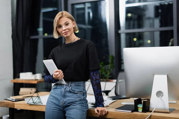 Cheerful blonde woman in glasses holding digital tablet near desk in office — Stock Photo