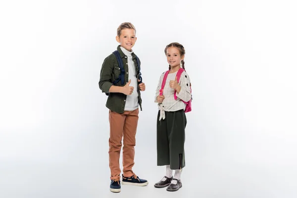Cheerful schoolkids standing with backpacks and showing thumbs up on white — Stock Photo