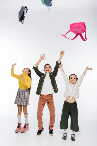 Cheerful schoolboy and schoolgirls throwing backpacks on white — Stock Photo