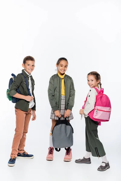 Happy schoolboy and cheerful schoolgirls standing with backpacks on white — Stock Photo