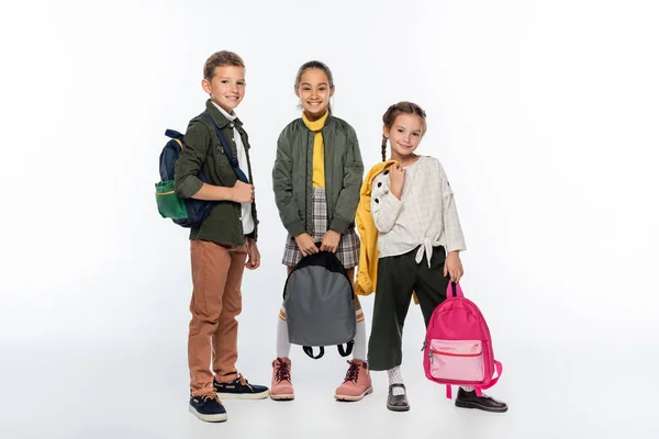 Cheerful schoolboy and positive schoolgirls standing with backpacks on white — Stock Photo