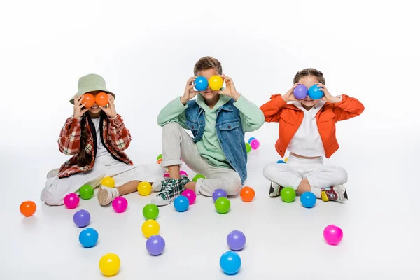 Niños preadolescentes felices sentados y cubriendo los ojos con bolas de colores en el aire en blanco - foto de stock