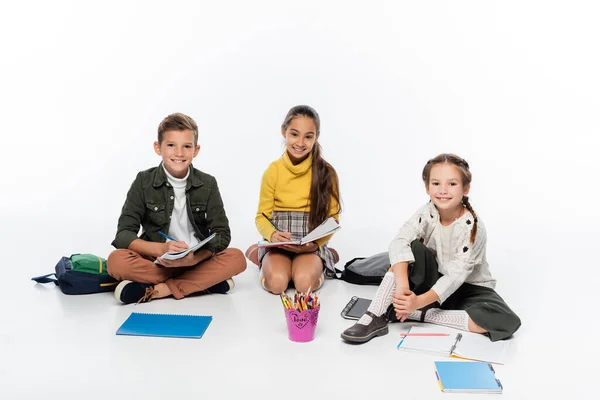 Alegre colegial y colegialas sentado cerca de mochilas y cuadernos mientras dibuja en blanco - foto de stock