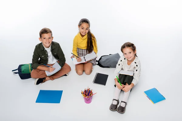 High angle view of cheerful schoolboy and schoolgirls sitting with notebooks while drawing on white — Stock Photo