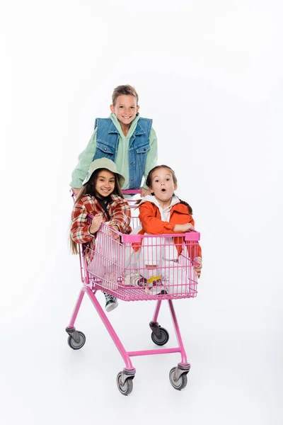 Comprimento total de menino feliz em pé atrás de meninas no carrinho de compras isolado no branco — Fotografia de Stock