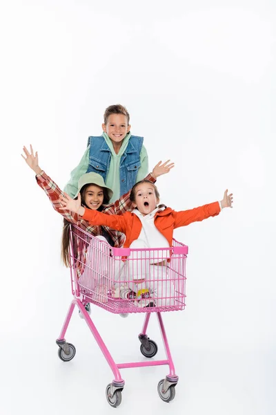 Happy boy standing behind amazed girls in shopping cart isolated on white — Stock Photo