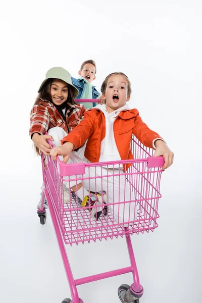 Fröhliche Kinder reiten in rosa Einkaufswagen isoliert auf weiß — Stockfoto