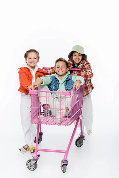 Niño feliz en el carrito de compras cerca de niñas alegres aislados en blanco - foto de stock