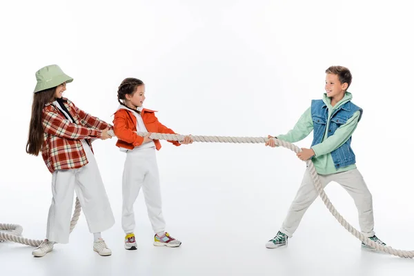 Vue de côté des filles gaies tirant la corde tout en jouant remorqueur de jeu de guerre avec garçon sur blanc — Photo de stock