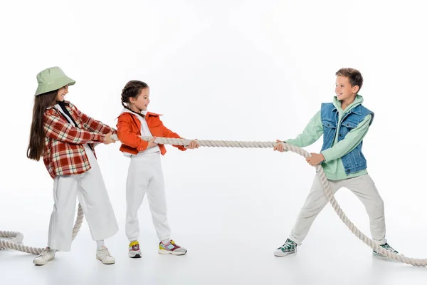 Side view of cheerful girls pulling rope while playing tug of war game with boy on white — Stock Photo