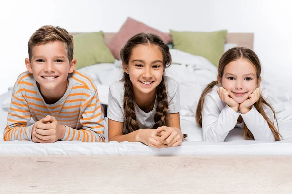 Niños alegres acostados en la cama y sonriendo aislados en blanco - foto de stock
