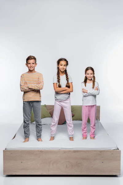 Cheerful kids standing with crossed arms on bed and smiling on white — Stock Photo
