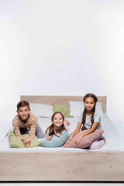 Crianças alegres sentadas na cama com travesseiros isolados no branco — Fotografia de Stock