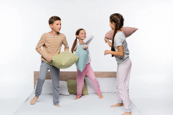 Joyful kids in pajamas having pillow fight while standing on bed isolated on white — Stock Photo