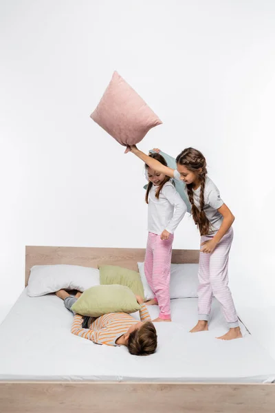 Веселі дівчата в піжамі мають постільну бійку, стоячи біля хлопчика на ліжку ізольовані на білому — стокове фото