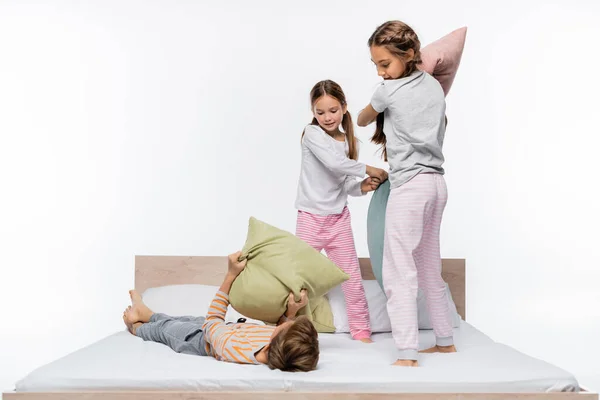 Feliz niñas en pijama teniendo almohada lucha mientras de pie cerca chico en cama aislado en blanco - foto de stock