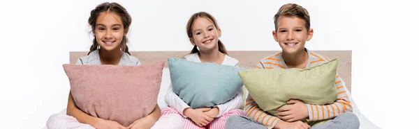 Fröhliche Kinder sitzen auf dem Bett und umarmen Kissen isoliert auf weiß, Banner — Stockfoto
