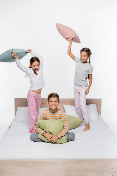 Alegre niñas en pijama teniendo almohada lucha con chico mientras de pie aislado en blanco - foto de stock