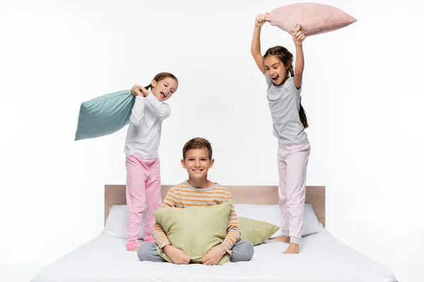 Heureux filles en pyjama ayant bataille d'oreillers avec garçon tout en se tenant isolé sur blanc — Photo de stock