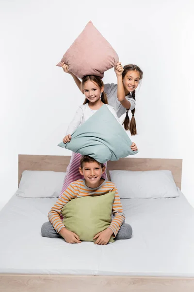 Playful girls in pajamas having pillow fight with boy while standing isolated on white — Stock Photo