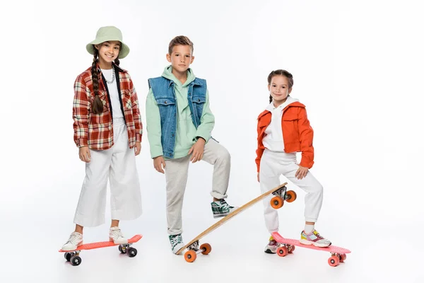 Longitud completa de los niños de moda sonriendo mientras está de pie con longboard y penny boards en blanco - foto de stock