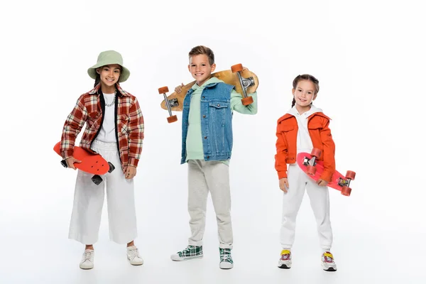 In voller Länge trendige Kinder lächeln, während sie Longboard und Penny-Boards auf weiß halten — Stockfoto