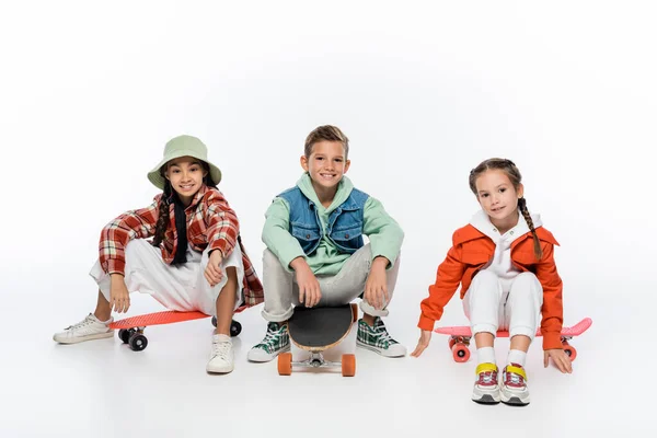 In voller Länge trendige Kinder lächeln, während sie auf Longboard und Penny Boards auf Weiß sitzen — Stockfoto