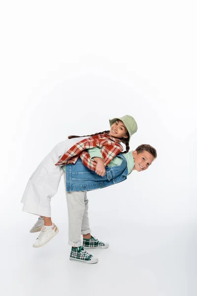 Full length of happy boy piggybacking cheerful girl in panama hat on white — Stock Photo