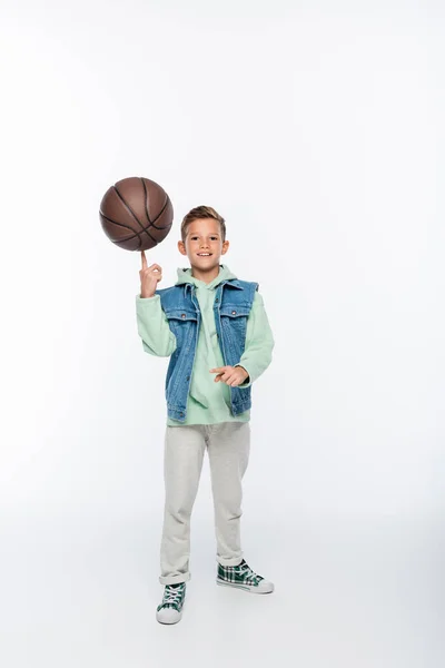 Повна довжина щасливого хлопчика, що крутить баскетбол на пальці на білому — стокове фото