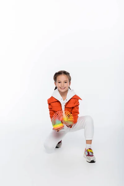 Lunghezza piena di bambino allegro in giacca arancione che gioca con arcobaleno slinky su bianco — Foto stock