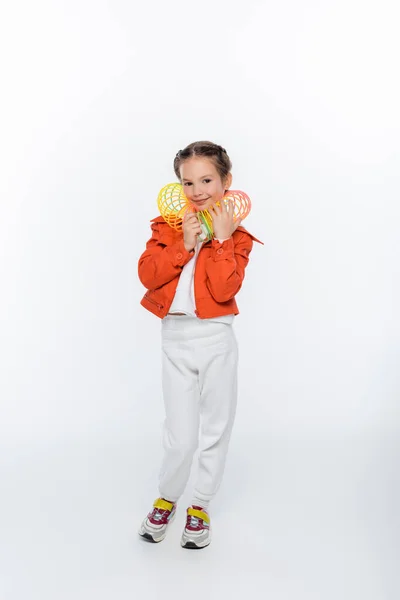 In voller Länge glückliches Kind in orangefarbener Jacke spielt mit Regenbogen slinky auf weiß — Stockfoto