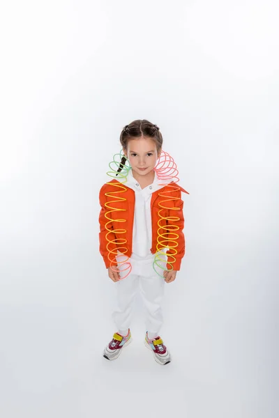 Longitud completa de niño sonriente en chaqueta naranja jugando con arco iris slinky en blanco - foto de stock