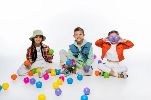 Crianças pré-adolescentes alegres sentadas perto de amigos cobrindo os olhos com bolas coloridas no branco — Fotografia de Stock