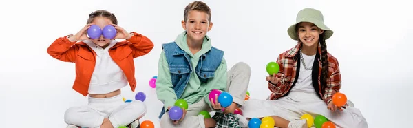 Cheerful preteen kids sitting near friend covering eyes with colorful balls on white, banner — Stock Photo