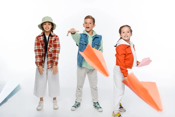 Longitud completa de niños preadolescentes felices lanzando aviones de papel en el aire en blanco - foto de stock