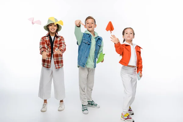 Longitud completa de niños preadolescentes alegres lanzando aviones de papel en el aire en blanco - foto de stock