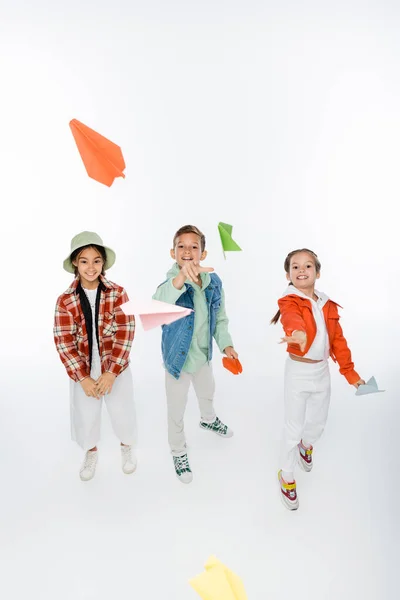 Longitud completa de los niños preadolescentes positivos lanzando aviones de papel en el aire en blanco - foto de stock