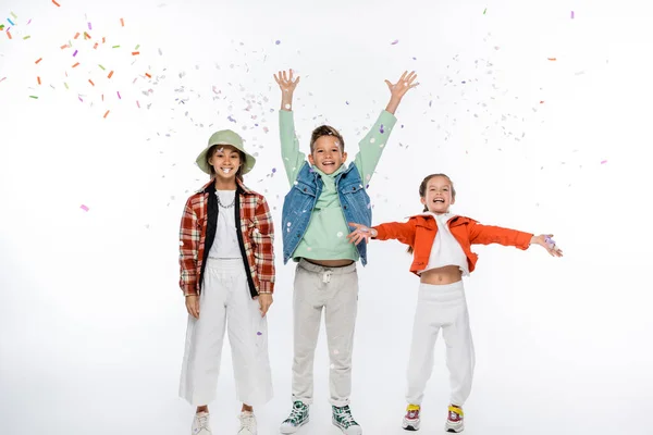 Full length of positive preteen boy with raised hands standing near girls and falling confetti on white — Stock Photo