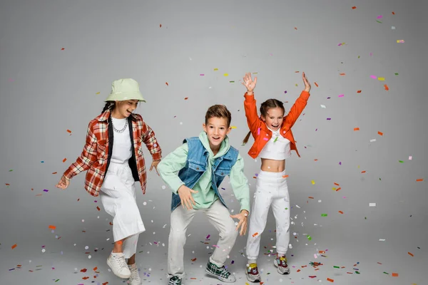 Full length of positive preteen kids standing near falling confetti on grey — Stock Photo