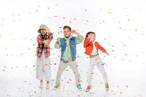 Pleine longueur de joyeux pré-adolescents enfants debout près de tomber confettis sur blanc — Photo de stock