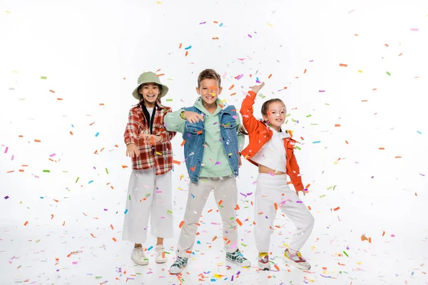 Pleine longueur de souriants préadolescents enfants debout près de tomber confettis sur blanc — Photo de stock