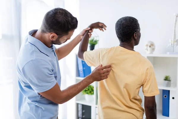 Junge Rehabilitologin untersucht Rücken eines afrikanisch-amerikanischen Patienten im Krankenhaus — Stockfoto