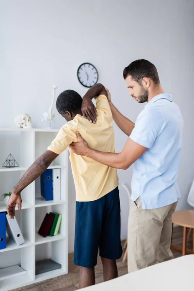 Joven médico tocando paciente afroamericano mientras lo diagnostica en centro de rehabilitación - foto de stock