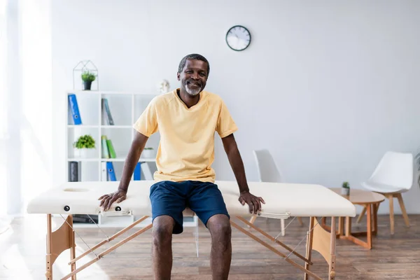 Uomo afroamericano di mezza età sorridente alla macchina fotografica mentre seduto sul lettino da massaggio nel centro di riabilitazione — Foto stock